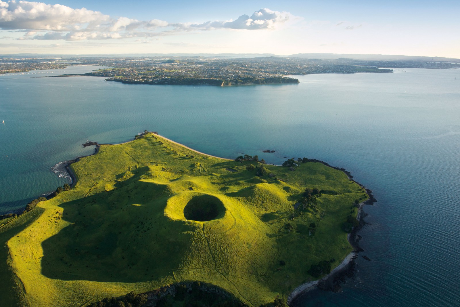 The verdant cone of Brown’s Island/Motokorea was the result of a cluster of volcanic events in the Auckland field beginning some 34,000 years ago. The lack of trees due to centuries of cultivation (it was one of the earliest European settlements in the Auckland area, purchased by William Brown and Logan Campbell in 1840), highlights the signatures of volcanic activity—a large scoria cone with a deep crater, a tuff ring forming the cliffs in the northeast, and the upper portions of lava flows that run into the sea.