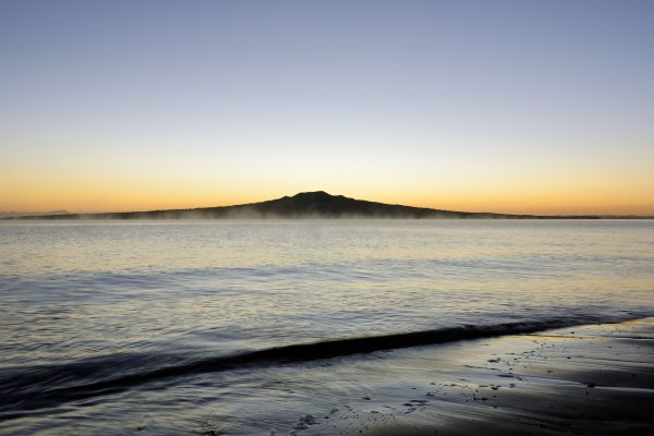 Rangitoto, wrote Ferdinand von Hochstetter in his 1864 Geologie von New-Seeland, “is for Auckland Harbour what Vesuvius is for the Bay of Naples, the ‘symbol’ of Auckland”. He described Rangitoto as small compared to the world’s more renowned shield volcanoes, but nevertheless “a true model of a volcanic mountain cone”. Seen here both softened by an early morning sea mist, and sharpened by the light of dawn, Rangitoto remains an enigma. It sits proudly offshore, and its exceptional symmetry affirms Auckland’s identity. Yet its size and eruptive history are exceptional too, and serve as a warning. Another Rangitoto in the wrong place could threaten the city’s population, infrastructure and identity.