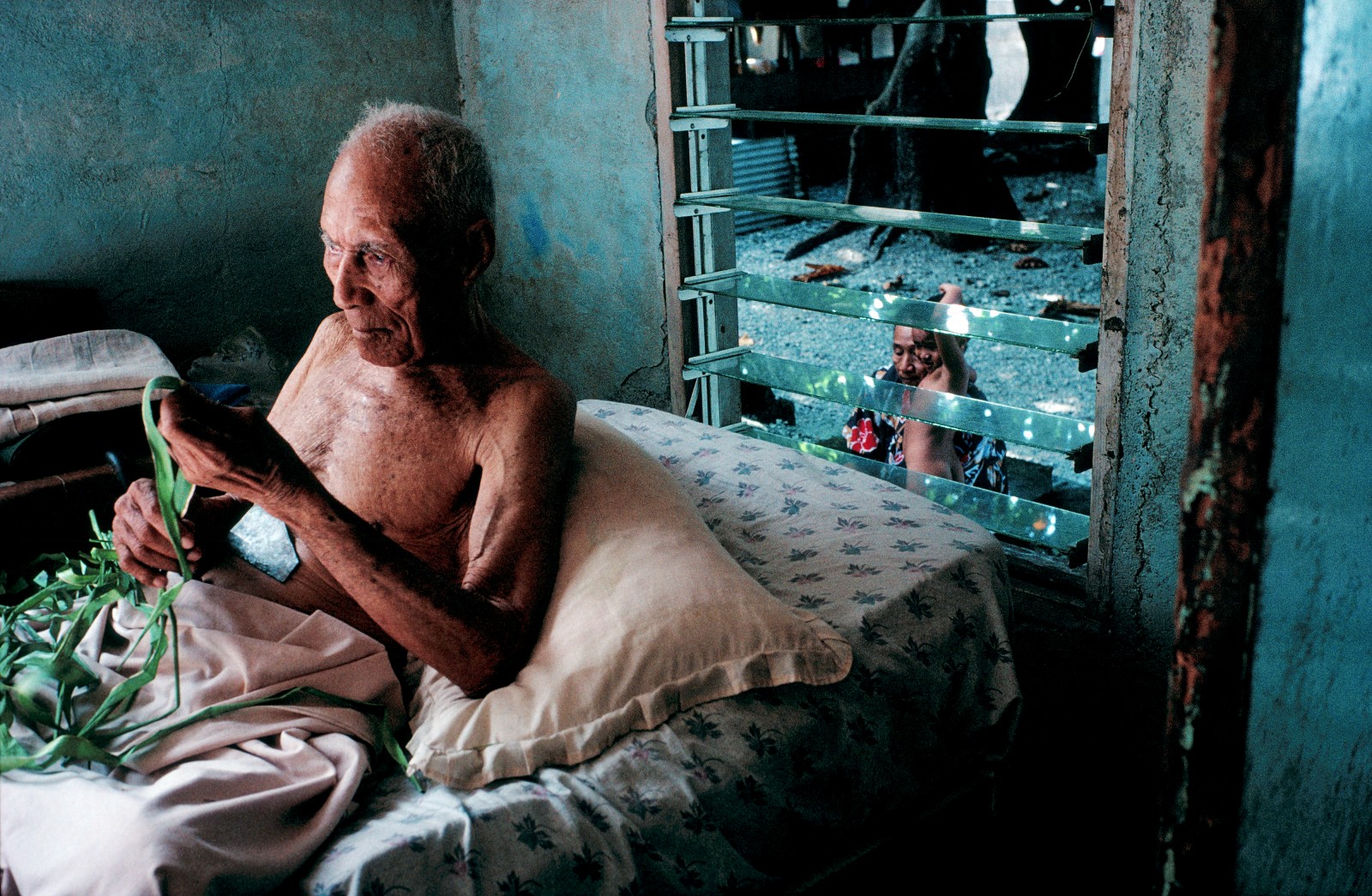 Heanoa Iha'ako is blind and deaf, but he's doing his bit for the household-dressing coconut fronds-and repaying the devotion of his daughter Hiniua, who bathes her son outside. In Tokelau it is an unwritten law that the family will always care for the parents.